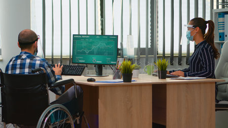 Woman entrepreneur bringing immobilized businessman at workplace with wheelchair wearing protection visor working in new normal business office. Businesspeople with masks respecting social distanceの写真素材