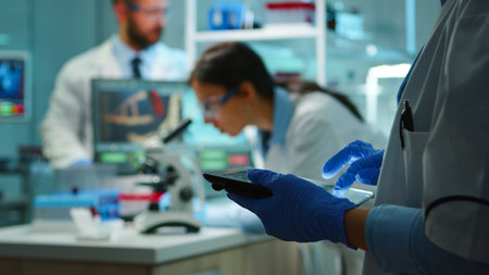 Close up of chemist nurse using tablet while team of scientists doing biological researches under microscope in backgorund late at night. Writing data into notepad in modern equipped laboratory.の写真素材