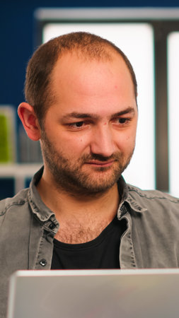 Enthusiastic business man writing a report sitting at table in modern startup business office sending mails typing on laptop, greeting colleague. Man working in financial company with diverse team.の写真素材