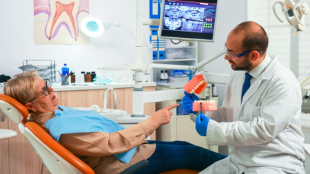 Young stomatologist explaining to senior patient procedure using model of dental teeth. Doctor holding sample of human jaw telling informations for keeping healthy teeth, digital x-ray in backgroundの写真素材