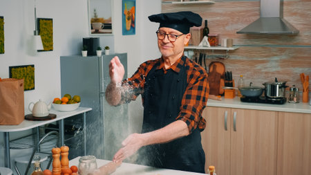 Senior chef playing with flour on the kitchen, clapping hands above the table. Professional cook...