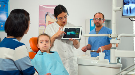 Stomatologist pointing on digital screen explaining x-ray to mother of girl patient. Doctor and nurse working together in stomatological clinic, examining, showing radiography of teeth on monitorの写真素材