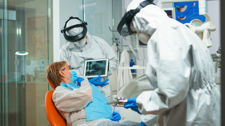 Stomatologist in protective suit reviewing x-ray of tooth with senior patient explaining treatment using tablet in covisd-19 pandemic. Medical team wearing face shield, coverall, mask and gloves.の写真素材