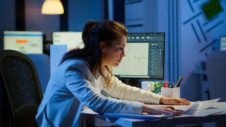 Focused woman architect checking and matching blueprints sitting at desk in front of computer late...
