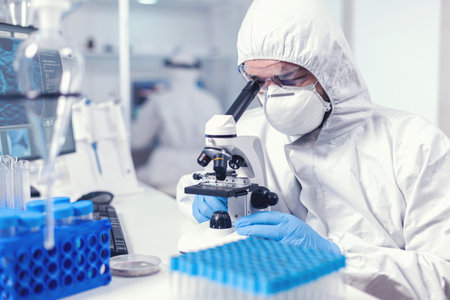 Focused healthcare researcher with face mask and ppe analysing covid using microscope. Scientist in...