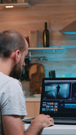 Freelancer editing a video using post production software working from home overtime sitting in the kitchen. Videographer processing audio film montage on professional laptop late at nightの写真素材