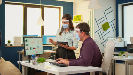 Business people wearing protective mask in new normal office making financial strategy pointing on desktop and taking notes on tablet. Multiethnic team working in company respecting social distance.の写真素材