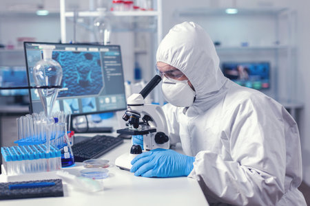 Healthcare researcher analying sample on microscope dressed in ppe suit. Virolog in coverall during...