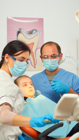 Little girl patient sitting in stomatological chair in dental office, visiting doctor for oral problems. Woman dentist with protective gloves and mask removes tooth decay drilling teeth during surgeryの写真素材