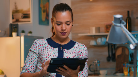 Businesswoman taking break using tablet late at night sitting in modern kitchen. Busy focused employee using modern technology network wireless doing overtime writing, searching.の写真素材