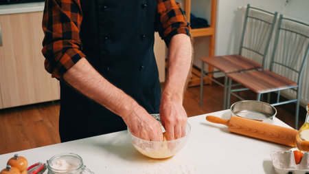 Elderly baker with kitchen apron adding flour on dough in glass bowl. Retired elderly chef with...