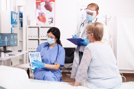 Nurse and doctor discussing with senior woman patient discussing treatmean holding tablet pc in time of covid19. Medical physician specialist during coronavirus outbreak discussing with patient.の写真素材