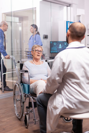 Paralyzed discussing with geriatrician sitting in wheelchair in the course of examination. Medical specialist wearing stethoscope discussing with invalid elderly woman in consultation room of private.の写真素材
