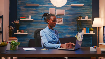 Focused african freelance writing on computer and using sticky notes for do list putting it on laptop sitting on desk in living room workplace late at night. Black woman working from home overtimeの写真素材