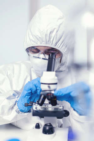 Medical engineer in ppe suit in modern laboratory looking at coronavirus sample using microscope....