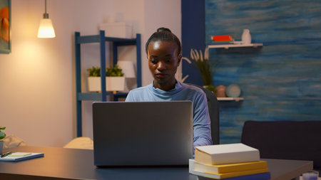 African woman coming in living room and typing on computer working from home overtime. Black entrepreneur sitting in personal workplace writing on keyboard looking at desktop respecting deadline.の写真素材