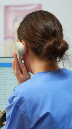 Orthodontist nurse talking at phone making dental appointments in modern equipped office, while specialist dentist with face mask examining patient with toothache sitting on stomatological chair.の写真素材