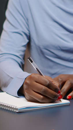 Close up of african businesswoman writing list of tasks for business project in notebook sitting at desk in living room working overtime. Black freelancer respecting deadline studying late at nightの写真素材