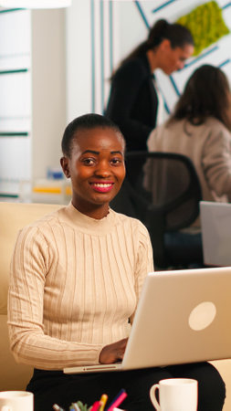 Portrait of african woman typing on laptop looking at camera smiling while diverse team working in background. Multiethnic coworkers talking about startup financial company in modern business officeの写真素材