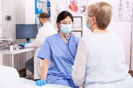 Nurse consulting senior patinet heart during health check up during covid19 outbreak. Medical specialist wearing face mask as safety precaution in the course of patient examination.の写真素材