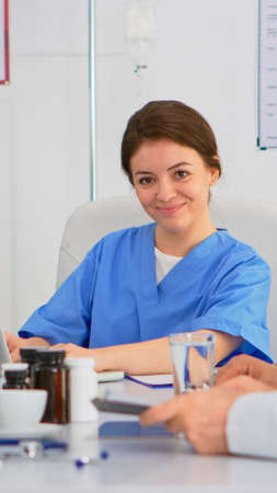 Nurse writing on laptop during conference and looking at camera smiling sitting on desk in hospital office, brainstorming room. Specialist woman in lab coat typing on notepad in medical clinic.の写真素材