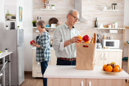 Senior couple arriving from supermarket with grocery bag and unpacking in kitchen early in the...