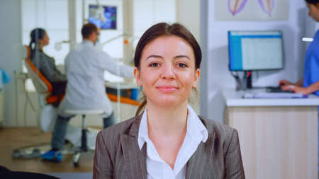 Portrait of smiling young patient looking on webcam sitting on chair in waiting room of stomatological clinic while doctor working in background. Stomatologist assistant typing on pc in dental officeの写真素材