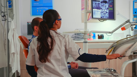 Dentist technician with glasses explaining the surgery pointing on monitor showing teeth implants. Orthodontist speaking to man with toothache sitting on stomatological chair.の写真素材