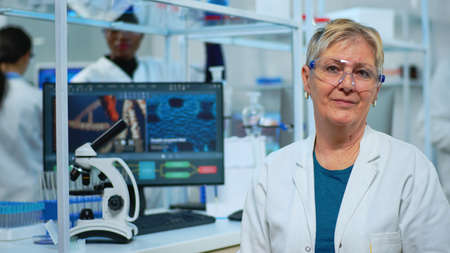 Portrait of old scientist woman smiling at camera in modern equipped lab. Multiethnic team examining...