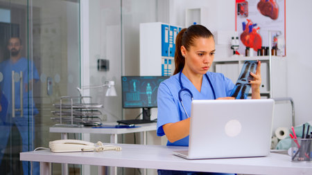 Medical woman nurse holding patient radiography typing on laptop sitting in hospital office. Radiologist in medical uniform looking at x-ray, examination, bone, practitioner, analyze, diagnosis.の写真素材