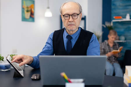 Portrait of mature man doing websurfing on laptop from home office. Elderly man entrepreneur in home workplace using portable computer sitting at desk while wife is reading a book sitting on sofa.の写真素材