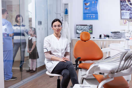 Ortodontist in dentistiry office sitting beside tools tray with assistant discussing with patietns in the background. Stomatolog in professioanl teeth clinic smiling wearing uniform looking at camera.の写真素材
