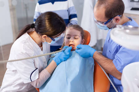 Little girl with mouth open in the course of cavity treatment sitting on dental chair. Mother with her kid in stomatology clinic for teeth examine using modern instruments.の写真素材