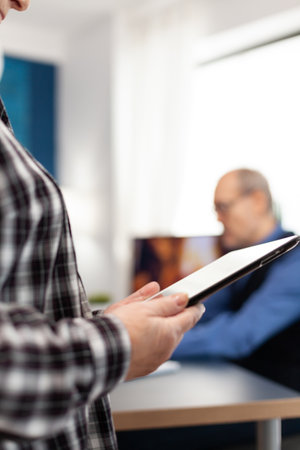 Midsection senior woman using digital tablet in home. Close up of elderly woman using moder technoloy tablet pc in home living room and husband working on laptop computer.の写真素材
