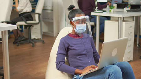 Black freelancer with protection mask against coronavirus sitting in middle of workplace office room...