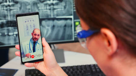 Close up of nurse talking on video call with specialist stomatologist using mobile phone sitting in modern dental clinic in front of pc with digital x-ray. Dentistry doctor explaining patient symptomsの写真素材