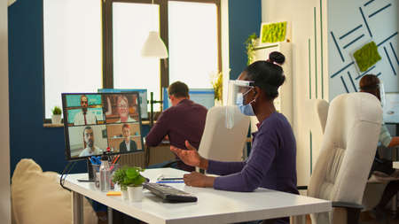 African businesswoman with face mask in office talking on video call with remote team during coronavirus epidemic. Having online conference, videomeeting colleagues working respecting social distance.の写真素材