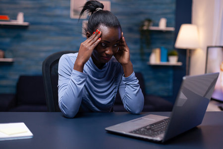 Dark skinned woman touching foarhead because of headache while working from home. Tired focused employee using modern technology network wireless doing overtime.の写真素材