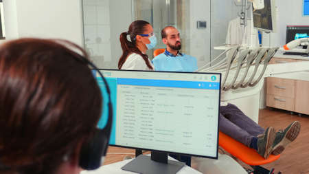 Orthodontist assistant making appointments using headset sitting in front on computer while doctor...