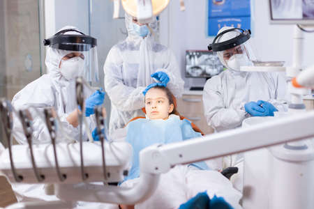 Mother dressed in ppe suit comforting daughter in the course of dental treatment. Stomatology team wearing ppe suit during covid19 doing procedure on child teeth.の写真素材