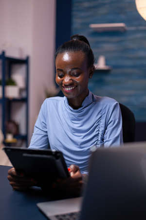 Smiling dark skinned woman browsing on tablet pc from home office late at night. Busy focused...