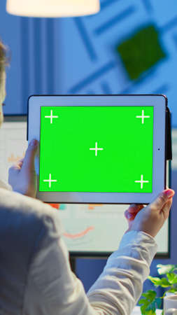 Businesswoman holding tablet with green screen monitor sitting at desk in business office late at...