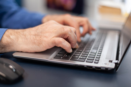 Close up of senior man hands typing on laptop keyboard. Elderly man entrepreneur in home workplace using portable computer sitting at desk while wife is holding tv remoteの写真素材