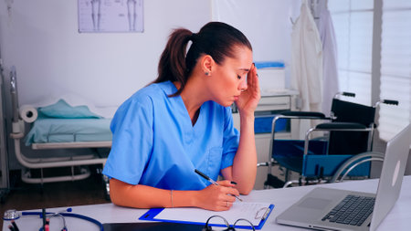 Overworked nurse wrinting on clipboard, working on laptop, taking off the glasses relaxing eyes. Medical physician in medicine uniform writing list of consulted, diagnosed patients, making research.の写真素材