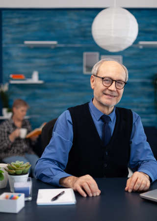 Cheerful senior businessman with gray hair. smiling at camera Elderly man entrepreneur in home workplace using portable computer sitting at desk while wife is reading a book sitting on sofa.の写真素材