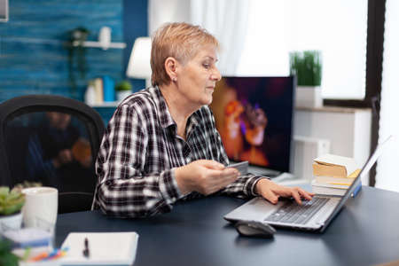 Positive senior woman paying bills holding plastic credit card. Joyful elderly woman using online banking for payment transcation surfing on internet from home living room.の写真素材