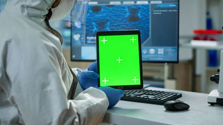 Chemist man wearing protection suit holding tablet with green mockup in modern equipped lab. Team of...