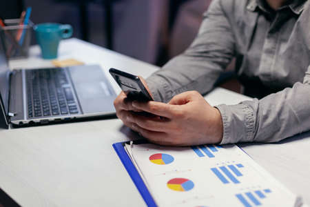 Close up of freelancer doing overhours texting on smartphone sitting workplace. Businessman using...