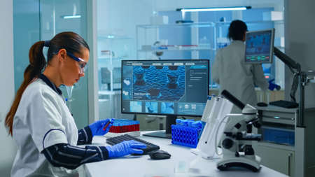 Chemist doctor holding test tube with blood sample entering datas in pc working in lab. Team of scientists analysing vaccine evolution using high tech researching diagnosis against covid19 virus.の写真素材