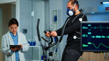 Monitor showing EKG reading of athlete running on cross trainer, while medical specialists supervise exercise, controlling physical activity, measuring heart rate in sports science laboratoryの写真素材